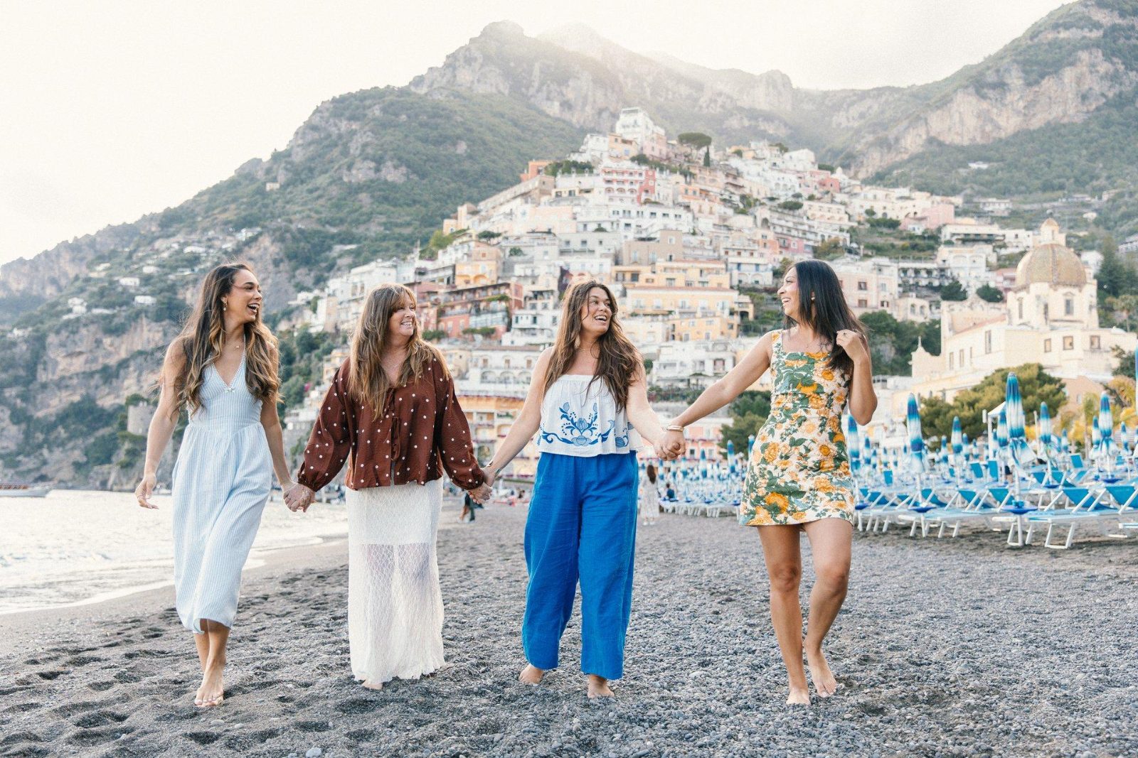 Four friends laughing and holding hands as they walk along the beach in Positano, with the colorful hillside town behind them